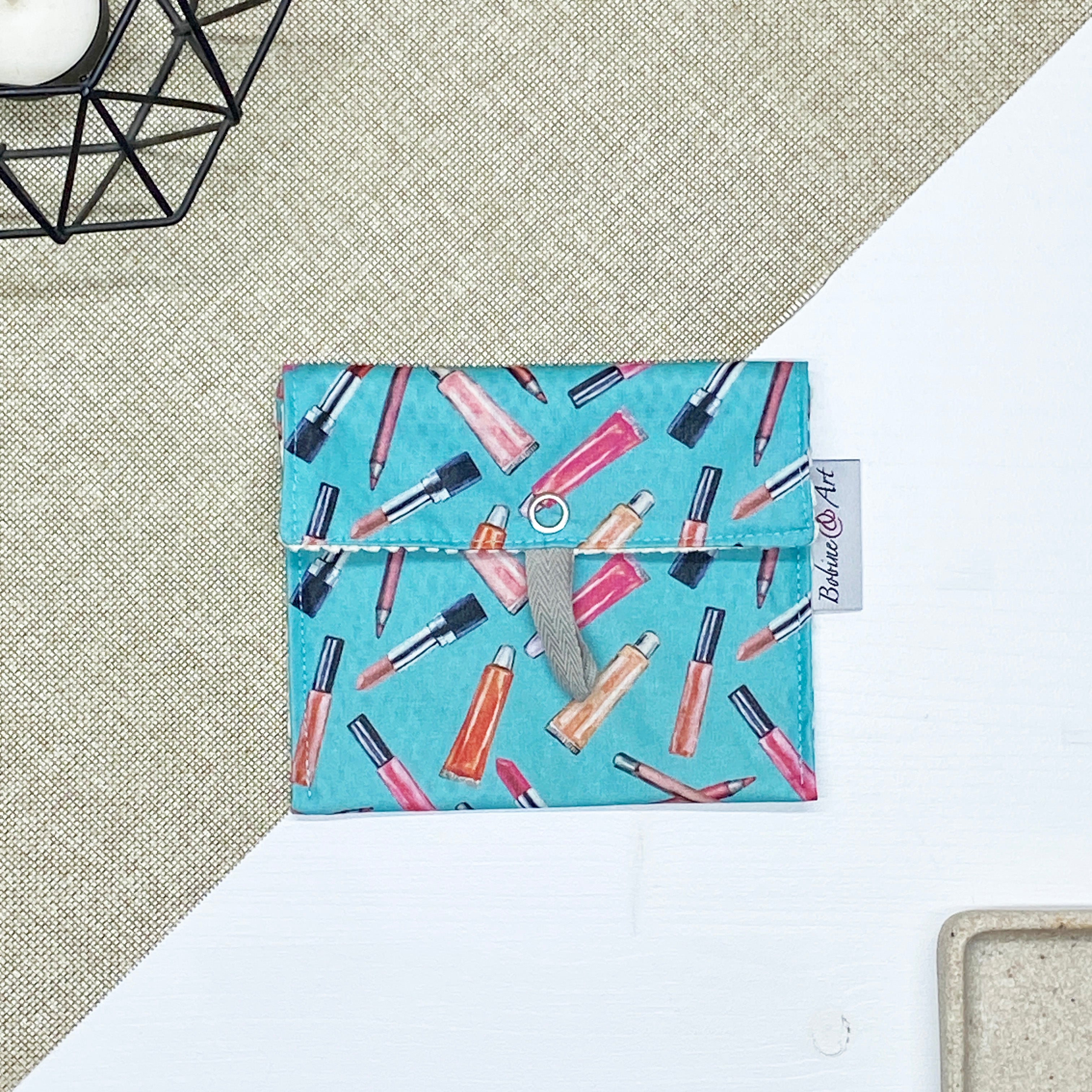 Porte-savon nomade imperméable fermé à motif maquillage avec rouges à lèvres, pinceaux et crayons sur fond bleu turquoise, format compact idéal pour transporter un savon solide lors des déplacements et voyages.