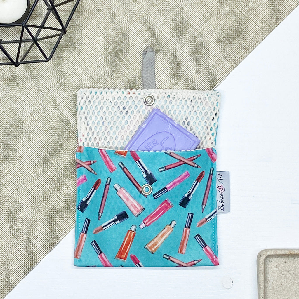 Porte-savon nomade imperméable à motif maquillage avec rouges à lèvres, pinceaux et crayons sur fond bleu turquoise, idéal pour transporter un savon solide ou des accessoires de toilette en déplacement.