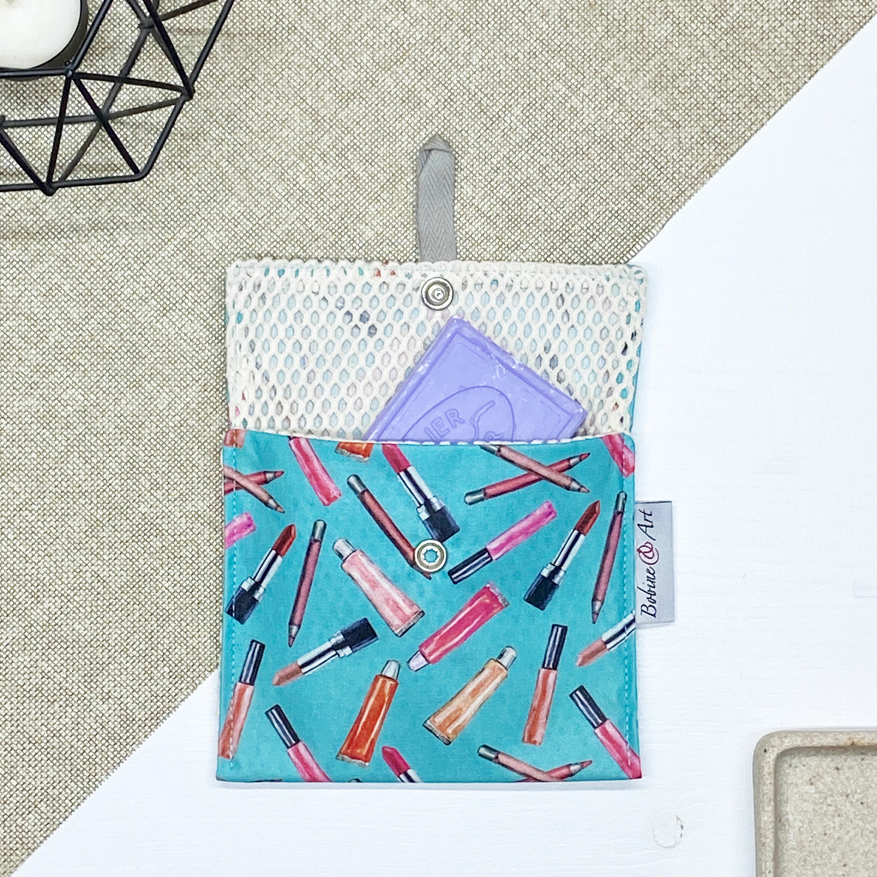 Porte-savon nomade imperméable à motif maquillage avec rouges à lèvres, pinceaux et crayons sur fond bleu turquoise, idéal pour transporter un savon solide ou des accessoires de toilette en déplacement.