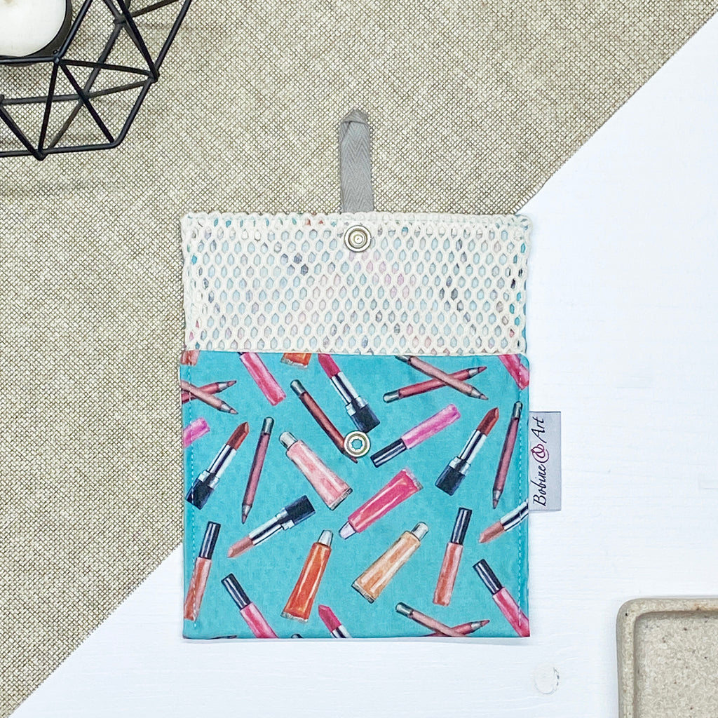 Porte-savon nomade imperméable ouvert avec filet respirant et motif maquillage composé de rouges à lèvres, pinceaux et crayons sur fond bleu turquoise, idéal pour transporter et faire sécher un savon solide en voyage.