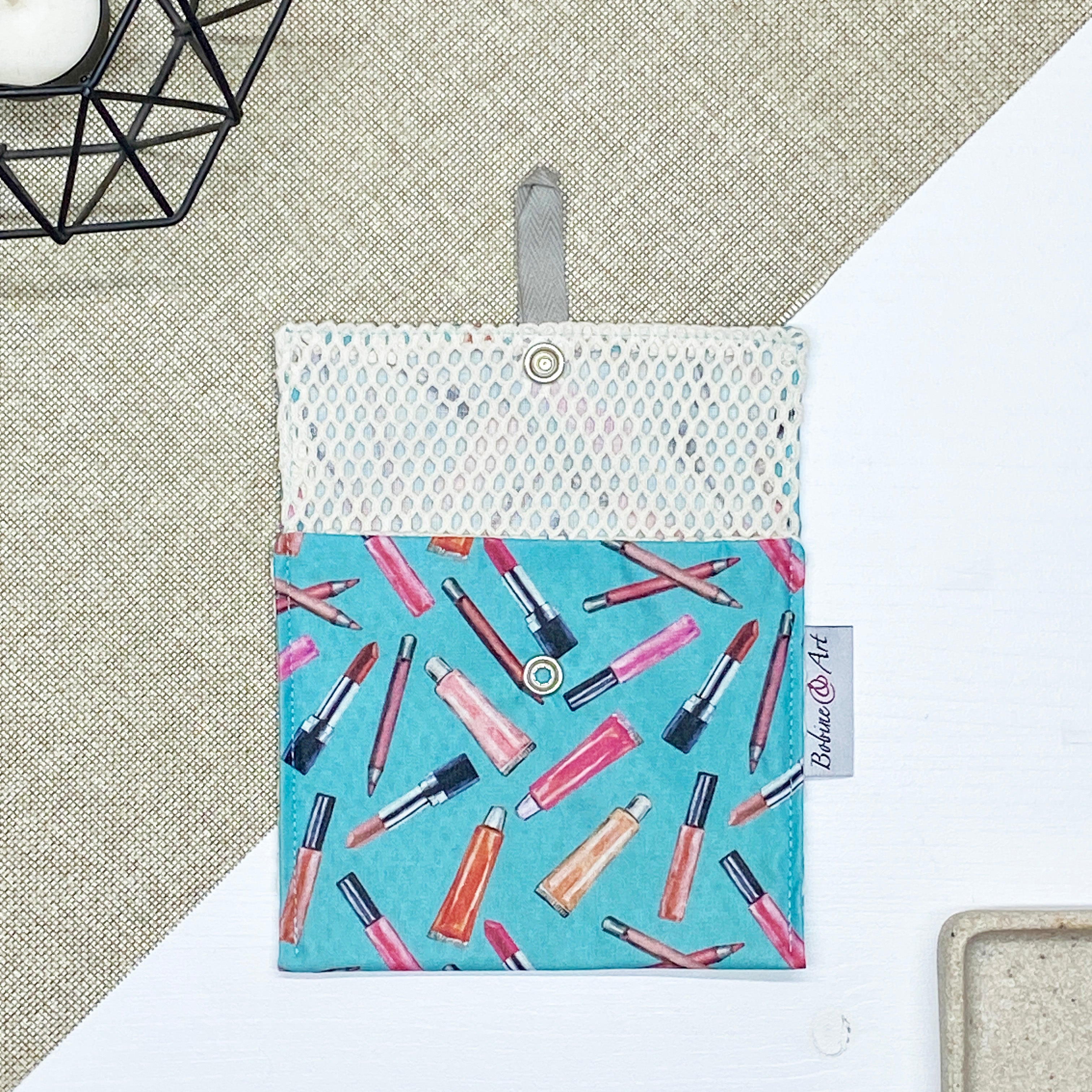 Porte-savon nomade imperméable ouvert avec filet respirant et motif maquillage composé de rouges à lèvres, pinceaux et crayons sur fond bleu turquoise, idéal pour transporter et faire sécher un savon solide en voyage.